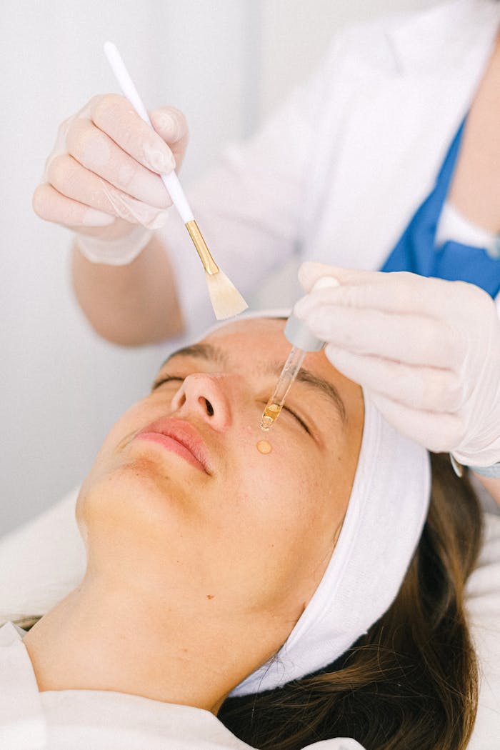 Skilled female beautician in uniform with brush applying moisturizing serum on beautiful female customer face skin in light modern beauty center
